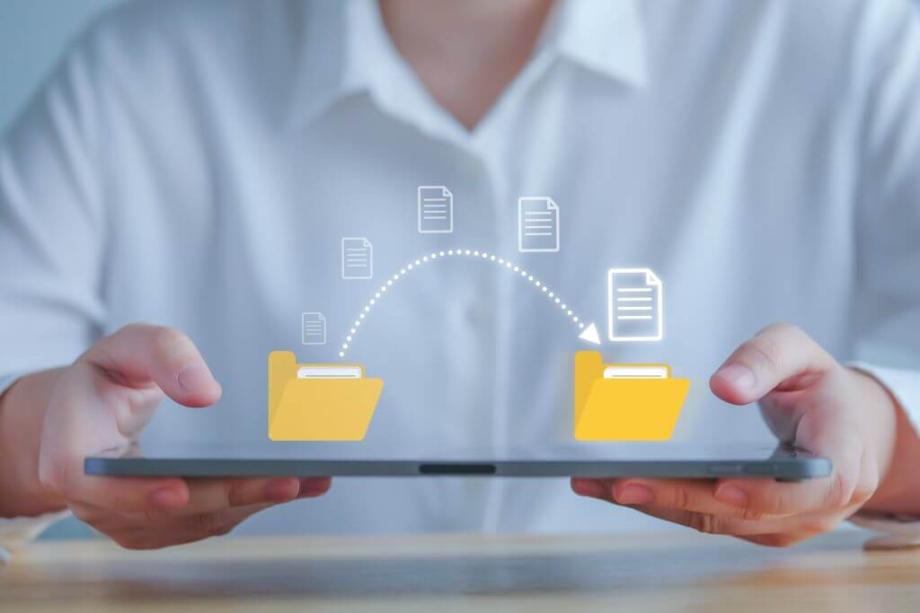 Zwei Händen halten Tablet, im Hintergrund sitzt ein Mann in einem weißen Hemd. Auf dem Laptop wird der Transfer von Daten grafisch dargestellt.