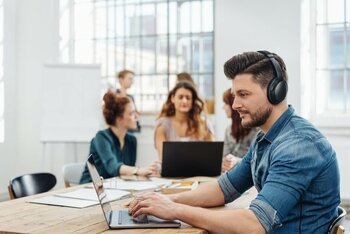Ein Mann mit Kopfhörern arbeitet an einem Laptop in einem Raum mit mehreren Personen.
