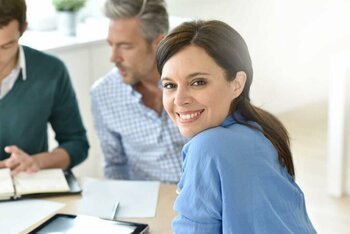 Eine lächelnde Frau blickt in die Kamera, im Hintergrund sitzen unscharf weitere Personen.