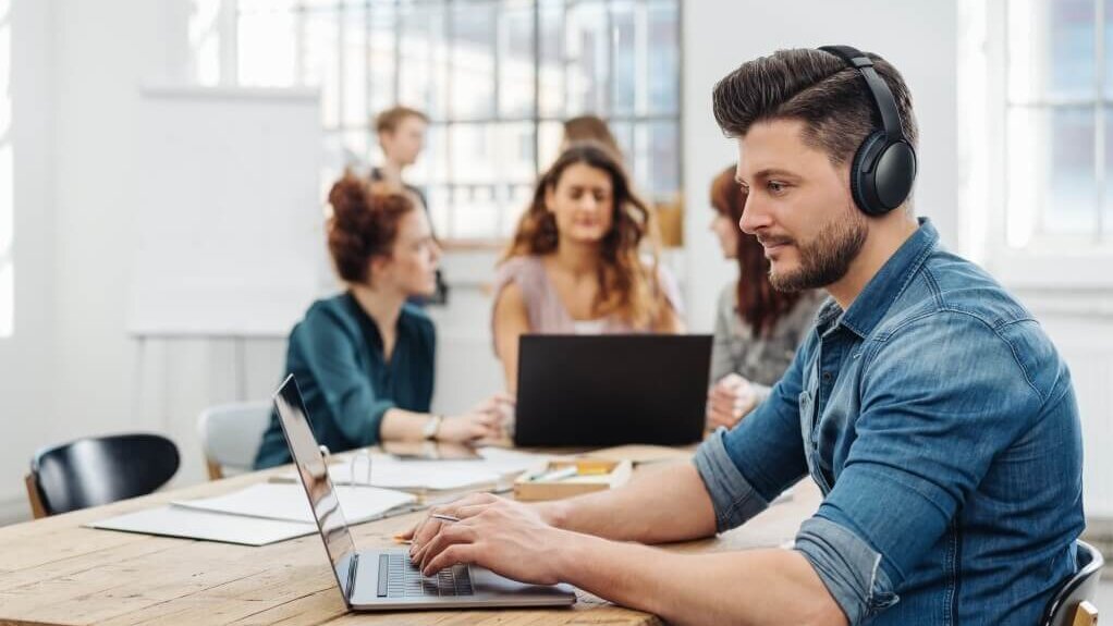 Ein Mann mit Kopfhörern arbeitet an einem Laptop in einem Raum mit mehreren Personen.
