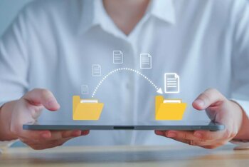 Zwei Händen halten Tablet, im Hintergrund sitzt ein Mann in einem weißen Hemd. Auf dem Laptop wird der Transfer von Daten grafisch dargestellt.