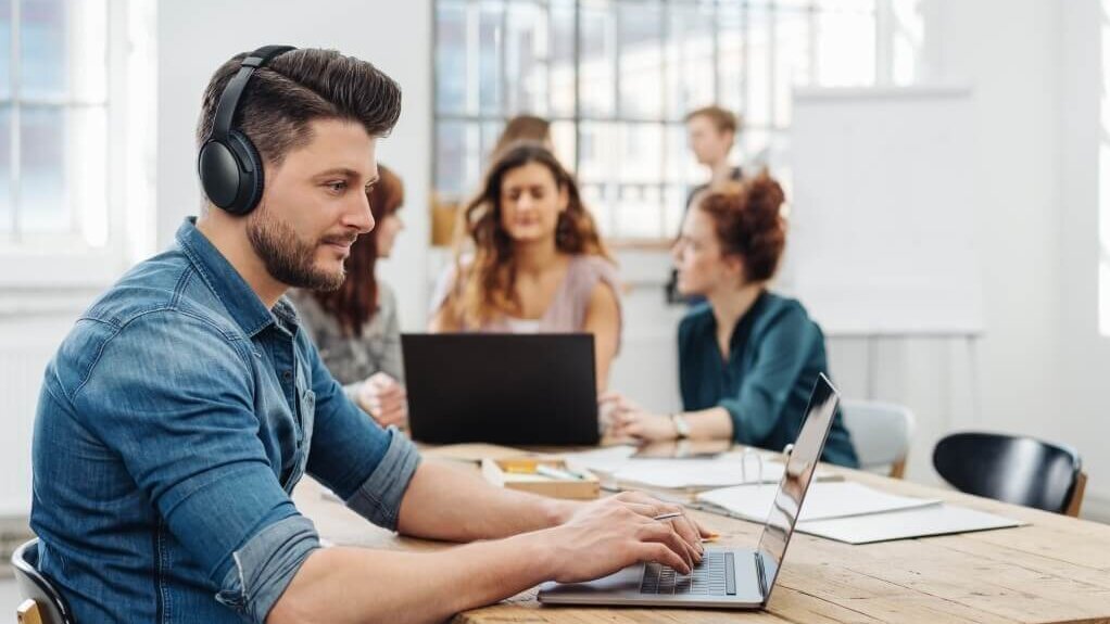 Ein Mann mit Kopfhörern arbeitet an einem Laptop in einem Raum mit mehreren Personen.