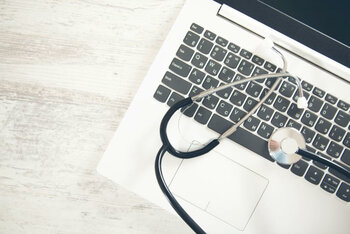Ein Stethoskop liegt auf der Tastatur eines silbernen Laptops auf einem Holztisch.